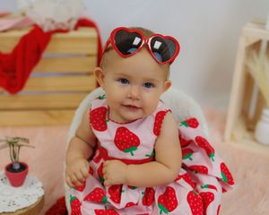 Baby wearing red and white strawberry print dress with matching bow headband poses cheerfully.