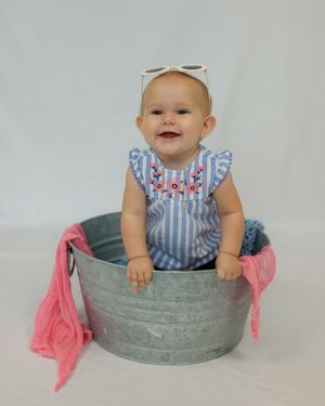 A happy baby wearing blue and white stripes sits in a rustic metal tub with pink fabric draped on the side.