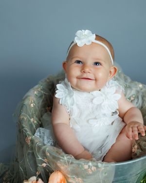 A baby wearing a white lace dress and floral headband sits in a decorative chair against a blue background.