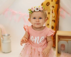 Baby wearing pink dress with 'one' text and floral crown headband poses during first birthday photoshoot.