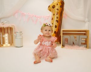 A baby wearing a pink tulle dress and bow sits on a wooden floor with a giraffe height chart in the background.