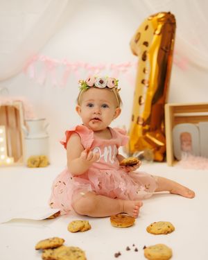 A baby in a pink tutu dress sits on a white backdrop with gold number one balloon and chocolate chip cookies.