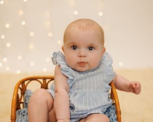 A baby wearing a light blue striped outfit sits in a wooden chair against a soft bokeh background.