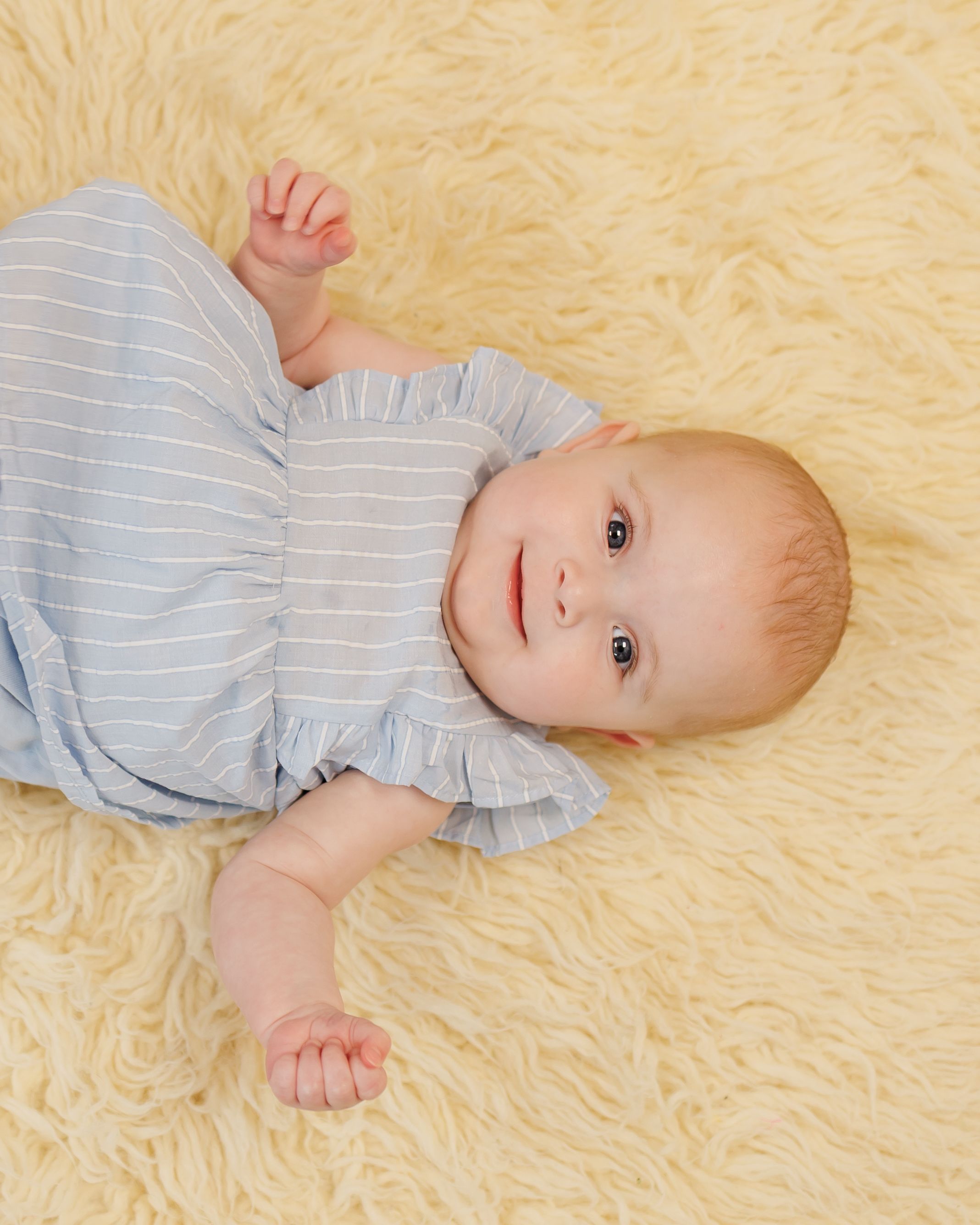 Small baby wearing a light blue striped outfit laying on soft cream-colored plush carpet.