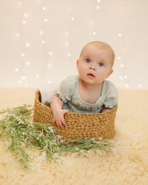 Baby wearing light blue dress sits in woven basket with greenery against beige backdrop with twinkling lights.