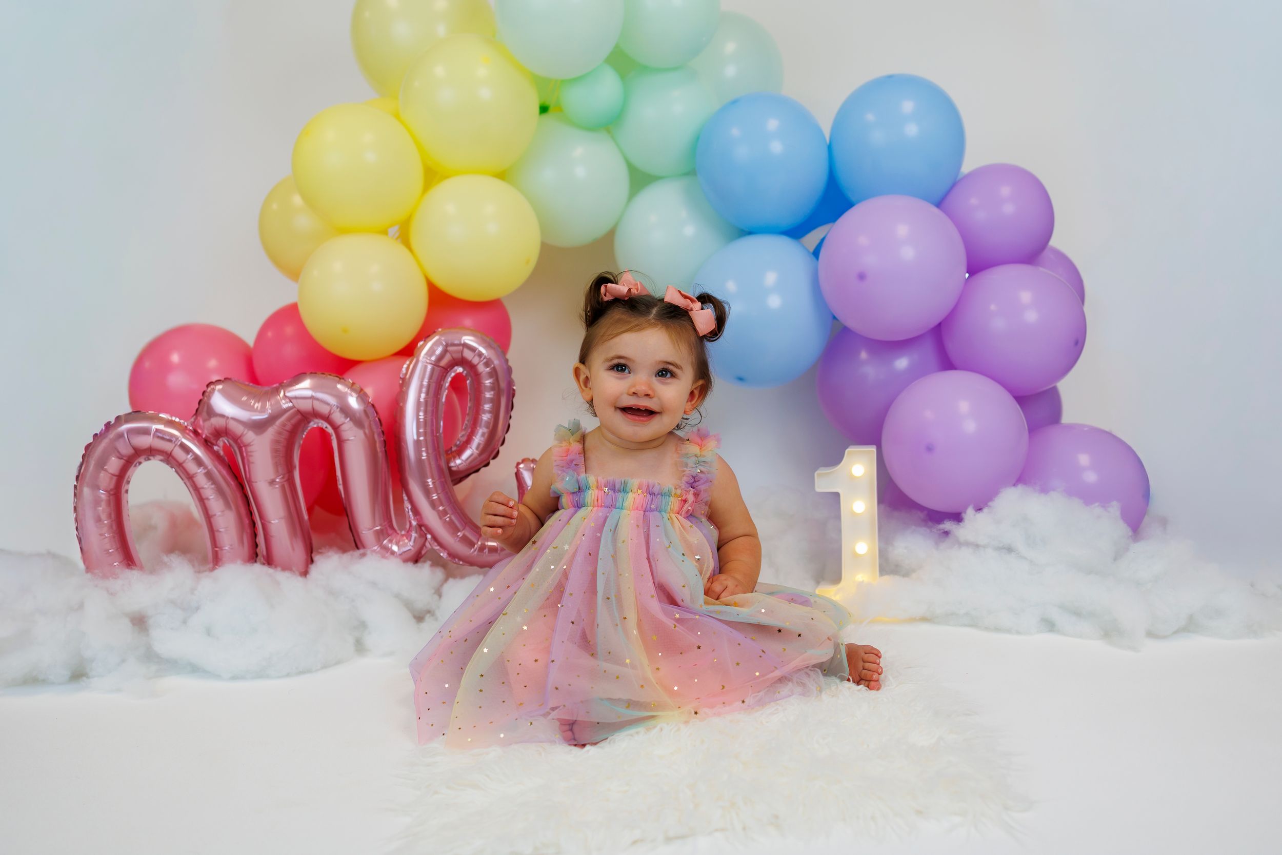 Rainbow balloon arch and pink number decorations create a festive backdrop for a cake smash photo session.