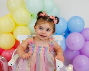 A baby in a pink floral dress smiles while surrounded by colorful pastel balloons at a birthday celebration.