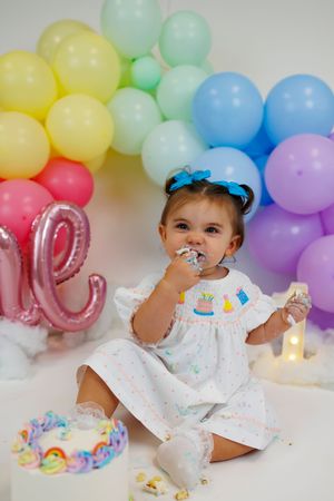 A colorful first birthday cake smash photo session with pastel balloon garland and pink number decorations in the background.