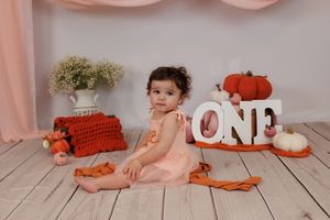 A baby in a pink dress sits on a wooden floor with pumpkins and fall decorations for a first birthday photoshoot.