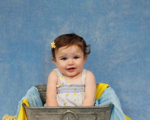 A baby wearing white sits in a rustic metal tub against a blue backdrop with yellow fabric accents.