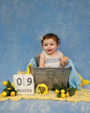 A baby in a vintage metal bucket with yellow tulips, blue backdrop, and blocks showing 9 month milestone.