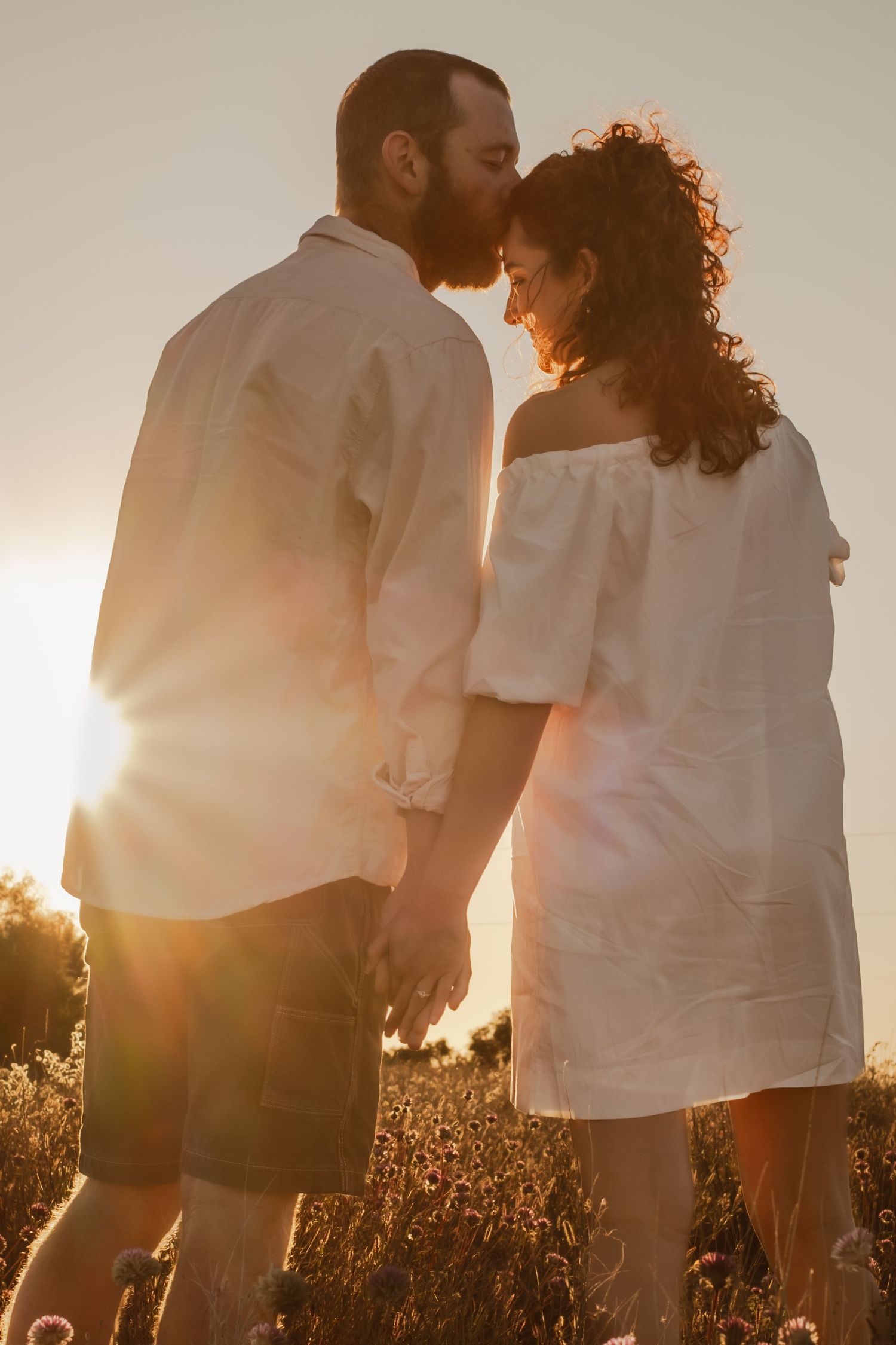 Couple in white clothing embracing in a sunlit field during golden hour.
