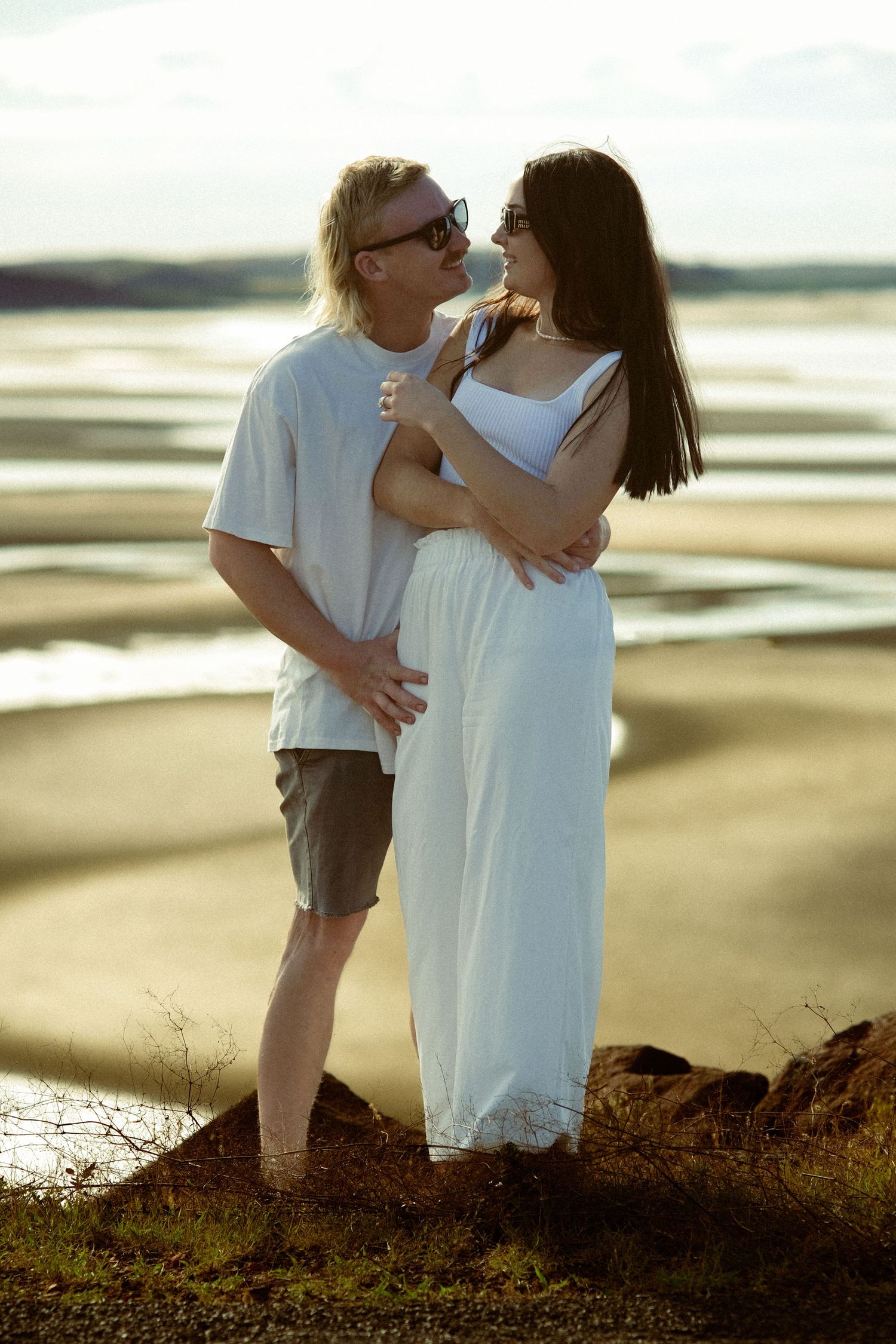 Couple in white clothing embrace on a coastal hill during golden hour.