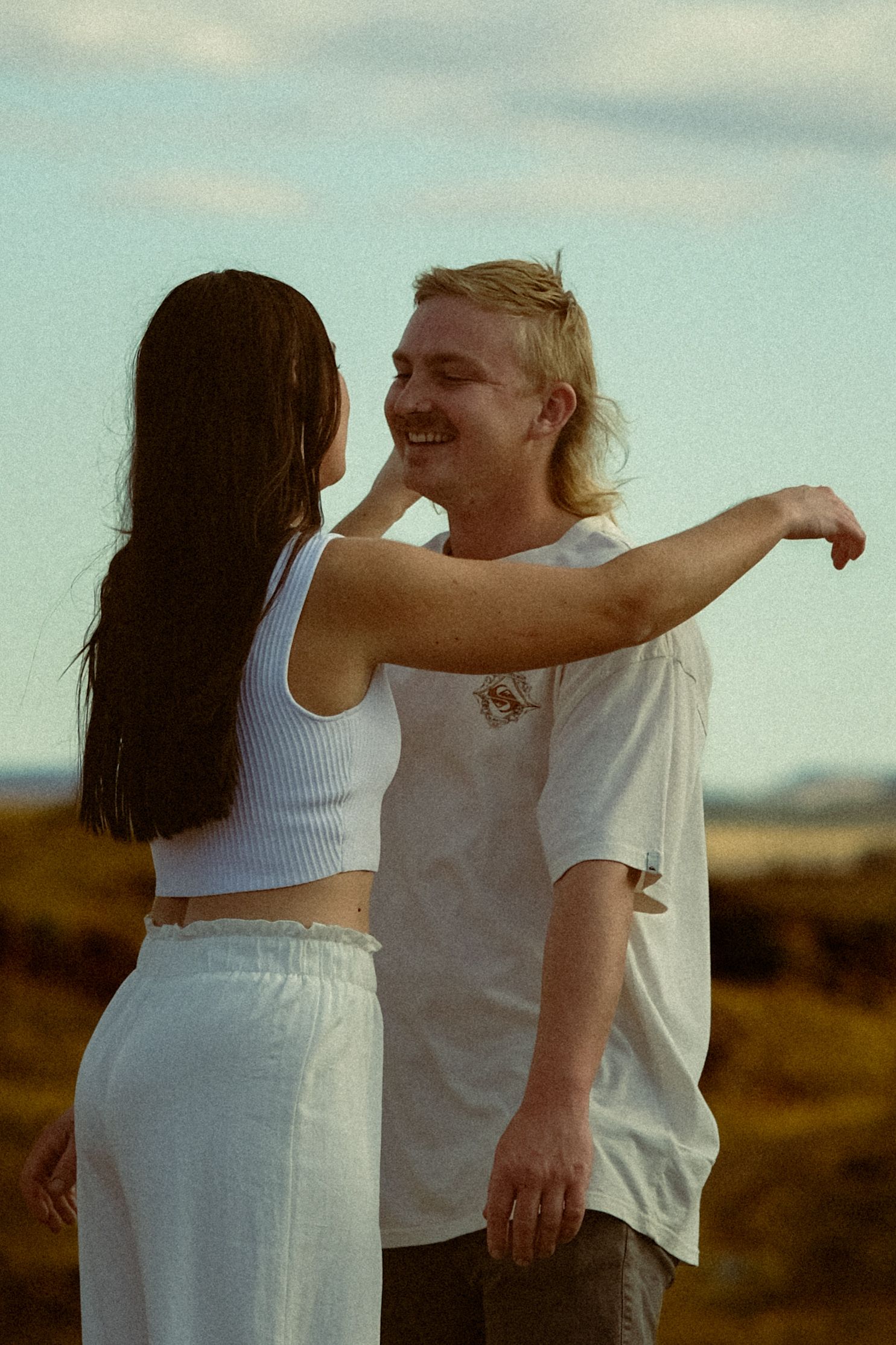 Couple in white clothing share playful moments against a soft sky backdrop.