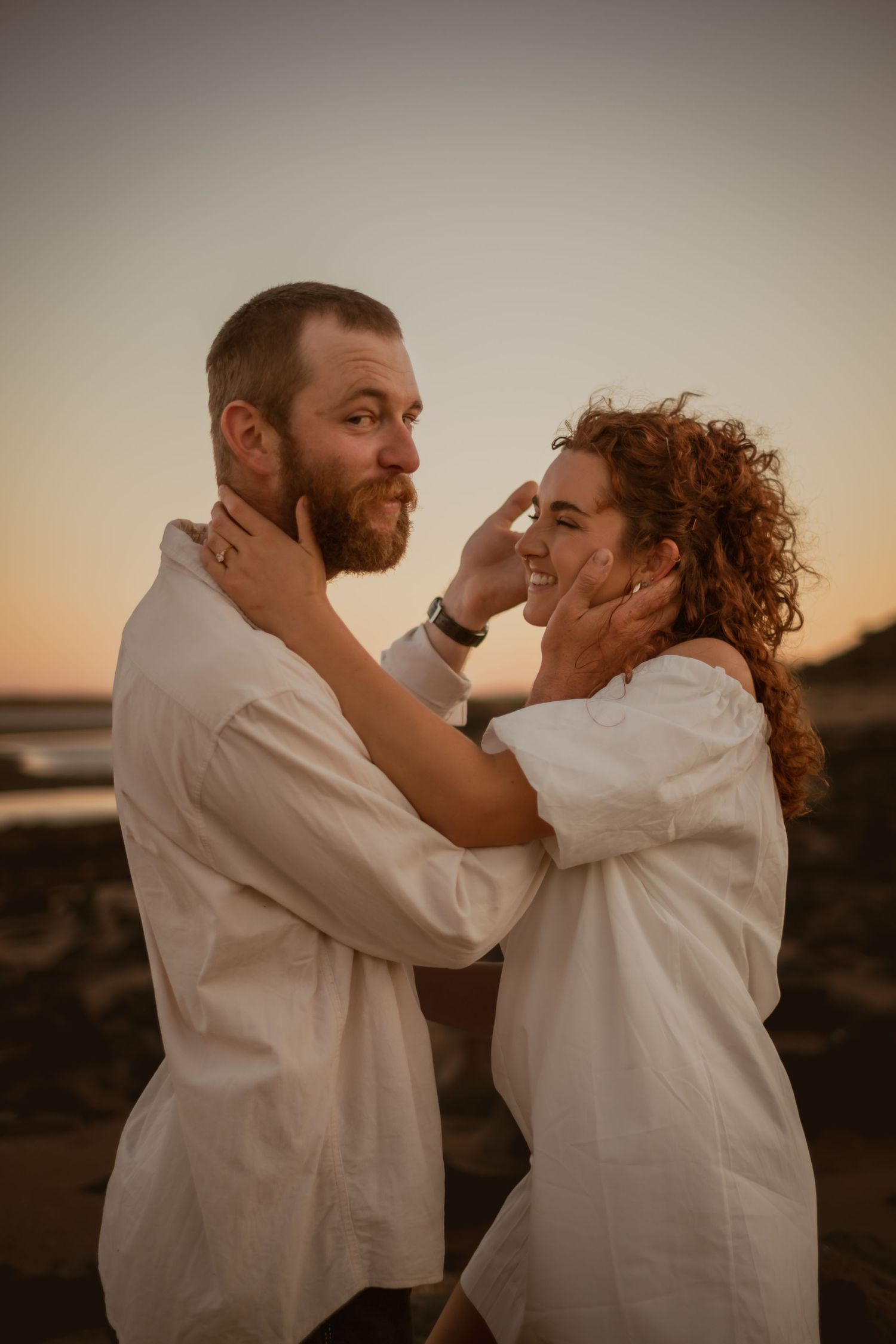 Couple in white shirts share an intimate moment during sunset in a rustic outdoor setting.