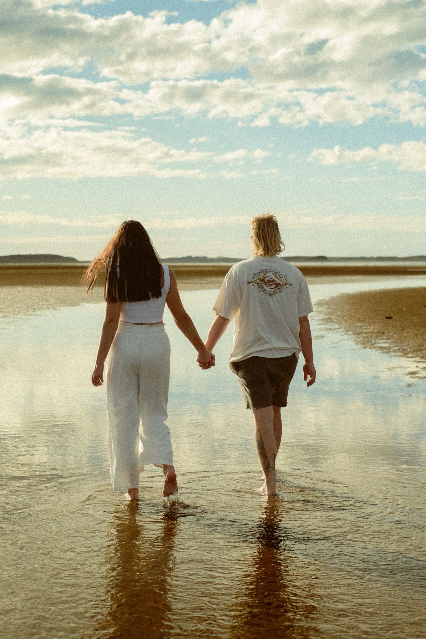 A couple walking hand in hand through shallow water on a beach at sunset.