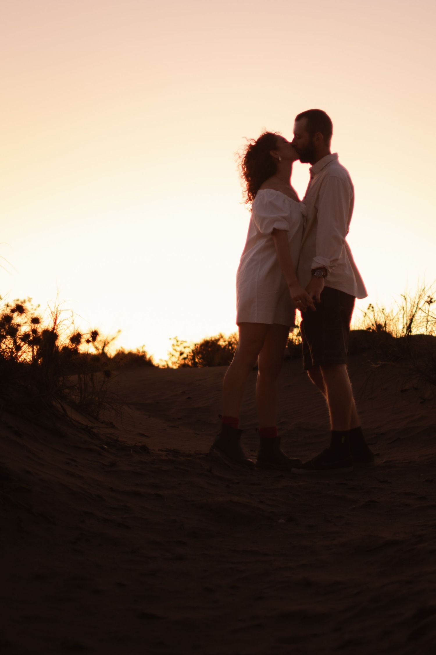 Romantic couple silhouetted against a warm sunset sky share an intimate moment on a hillside.