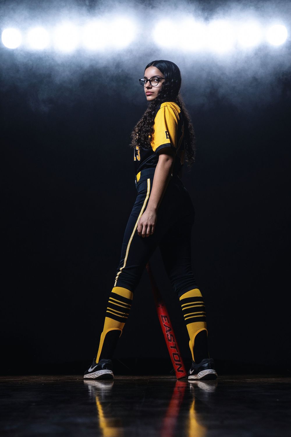 Athletic player in yellow and black uniform strikes dramatic pose on dark court with dramatic overhead lighting.