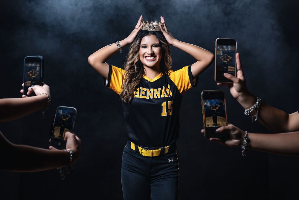 Student wearing university athletic jersey poses with crown while surrounded by phones taking pictures in dark studio setting.