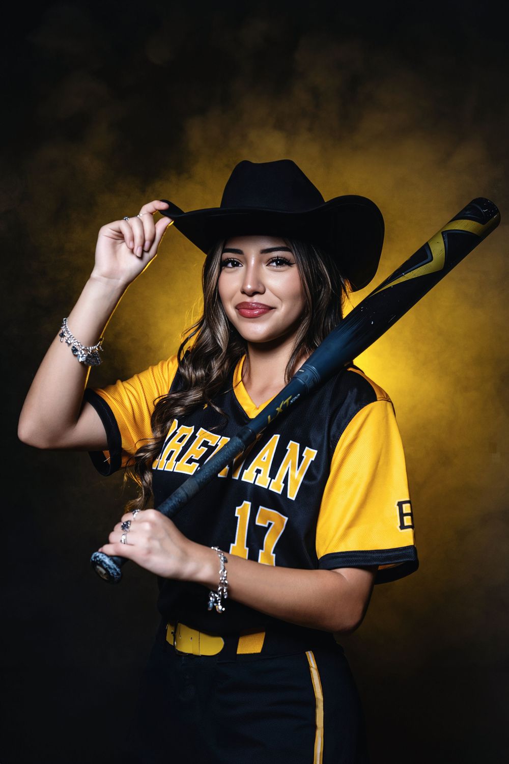Softball player in yellow Ethan jersey and black cowboy hat poses with bat against dark dramatic background.