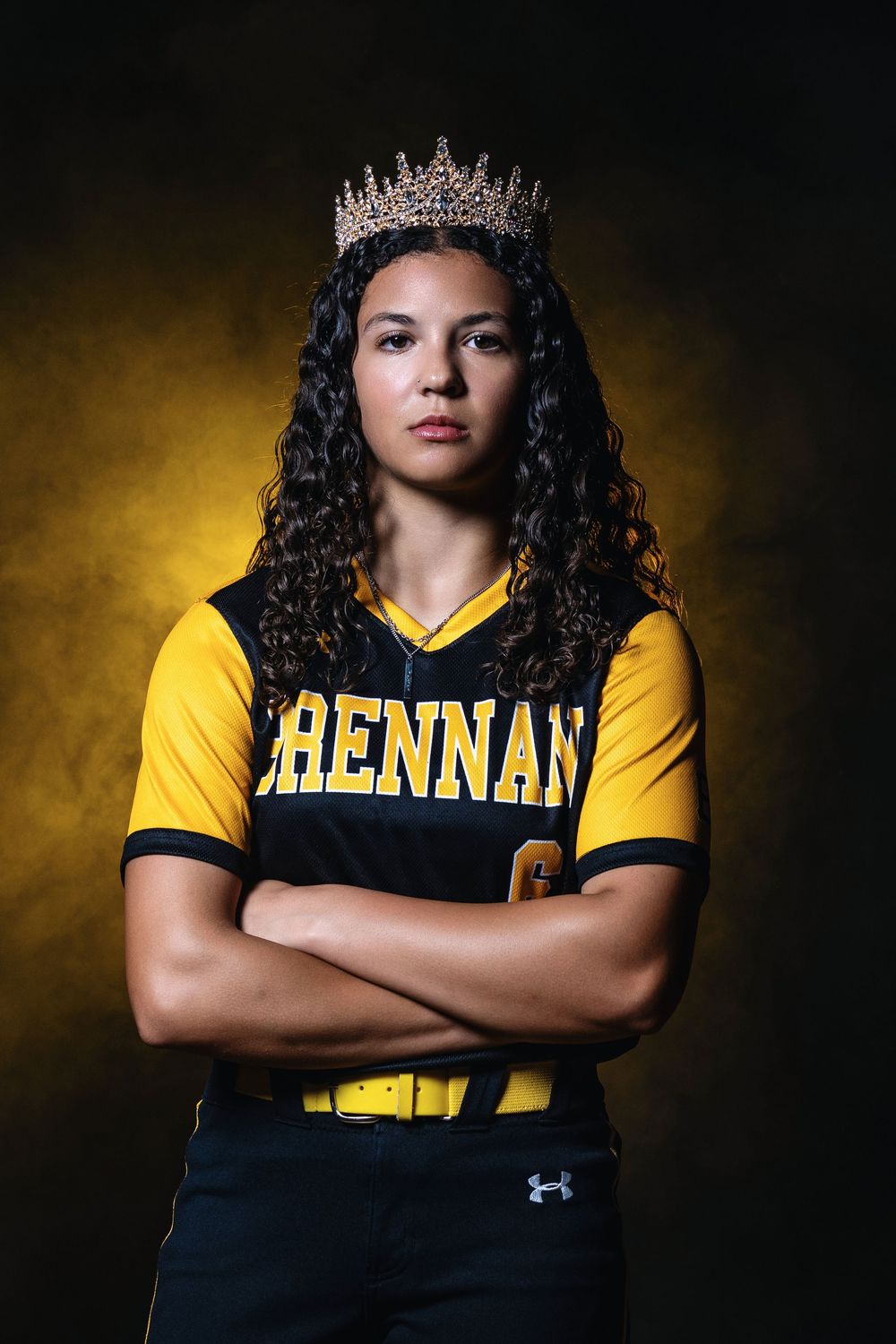 Student athlete wearing yellow Brennan team jersey and sparkling crown poses with crossed arms against dark background.