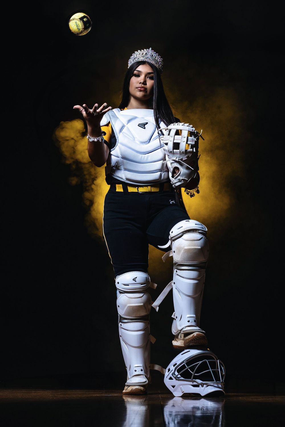 Softball player in protective gear poses dramatically with ball and glove against dark background with golden lighting effects.