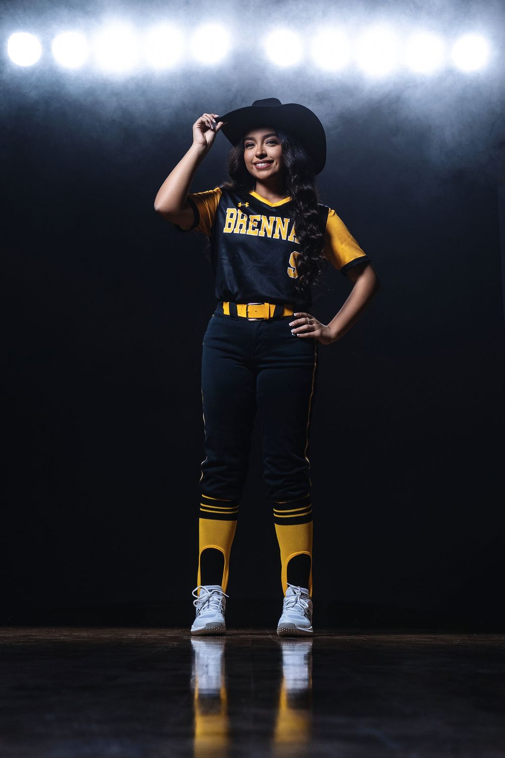 Athletic player in navy and gold softball uniform poses dramatically under stadium lights with a cowboy hat.