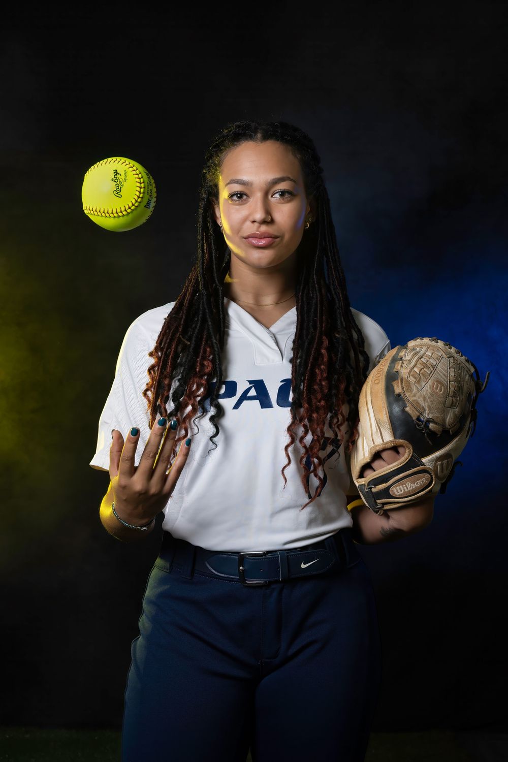 A softball athlete in a DePaul uniform poses with a glove and tossing a bright yellow softball against a dark background.