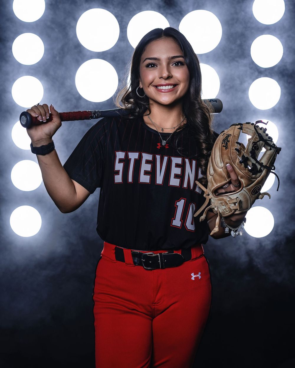 A softball player in a Stevens uniform #10 poses with a bat and glove against a backdrop of bright lights.