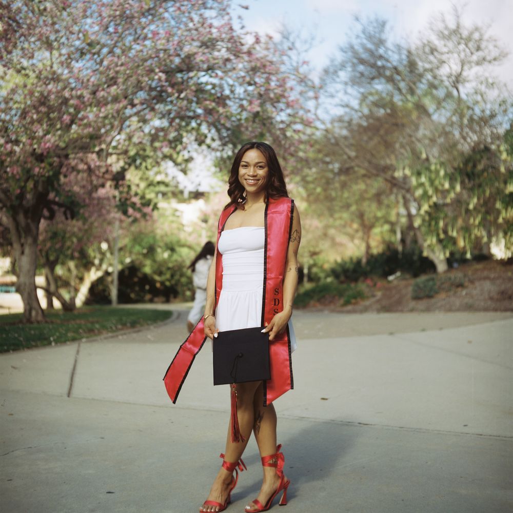 A young woman poses in front of the camera surrounded by trees with bright pink leaves