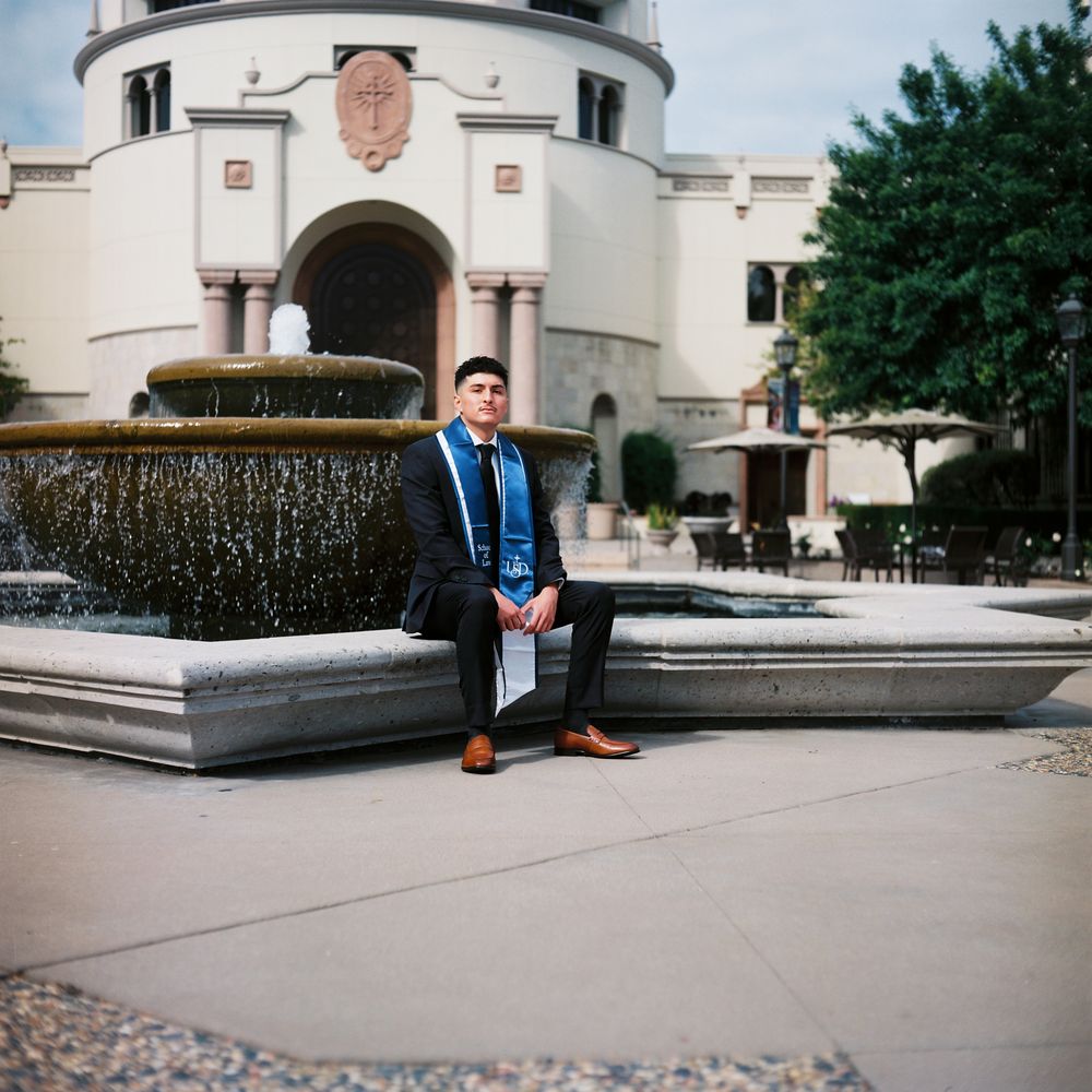 A young man sits down on the edge of a beautiful fountain facing the camera