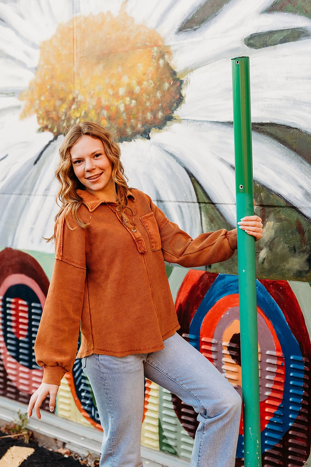 Figure stands in a brown knit sweater and jeans against colorful mural background with flower design overhead.