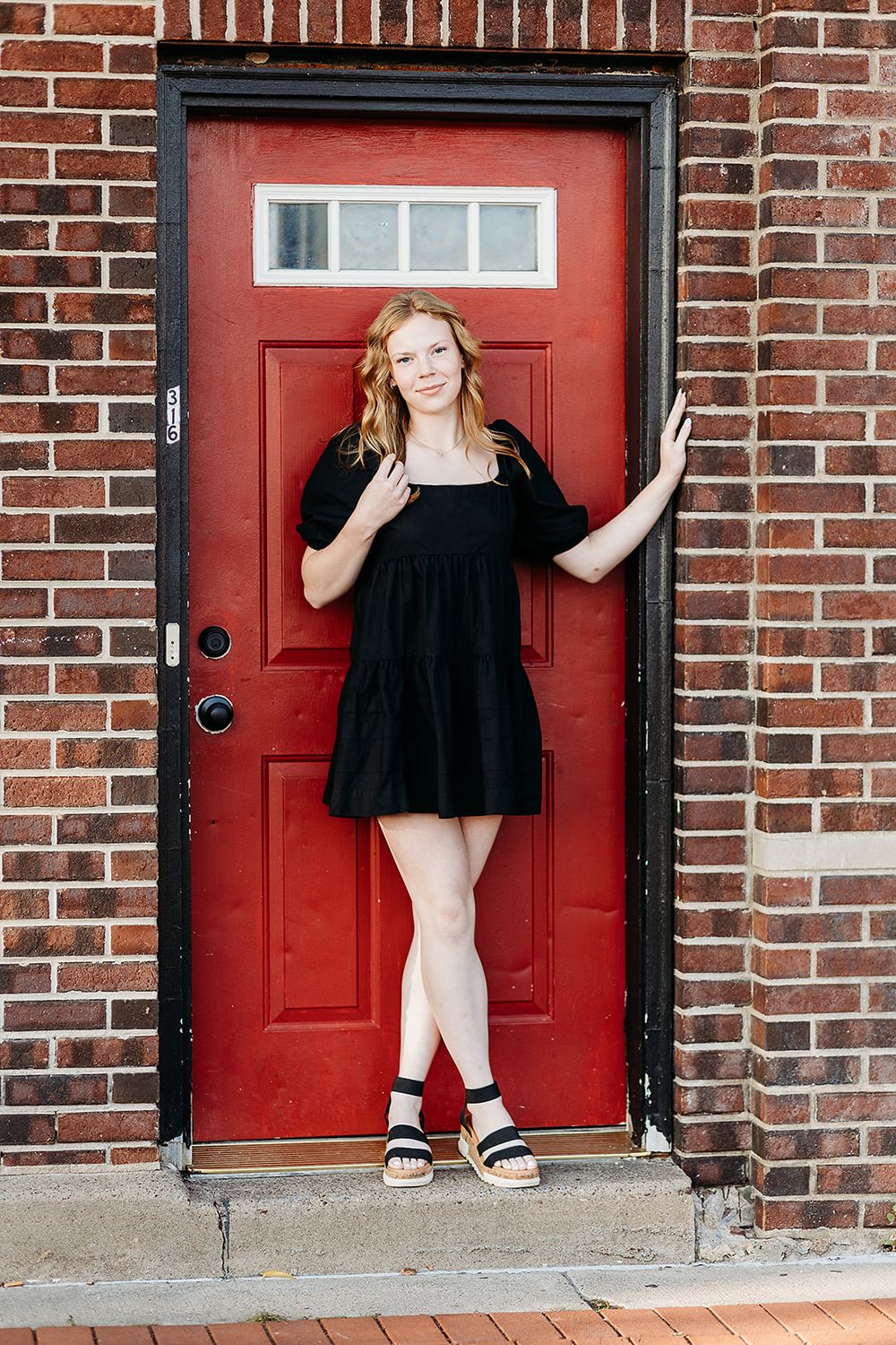 Person in black dress poses against vibrant red door with white transom window on brick building exterior.