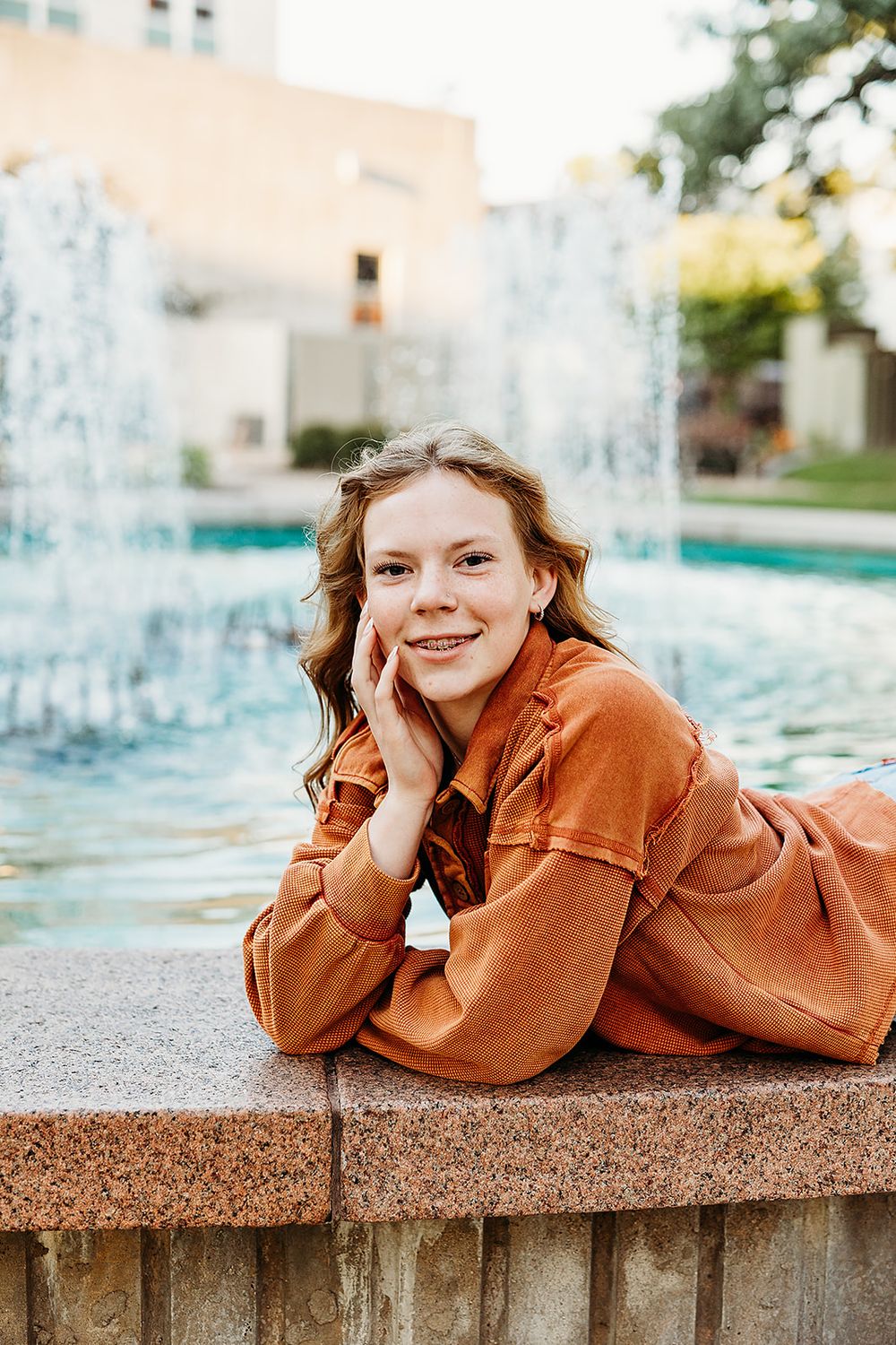 Person in burnt orange jumpsuit lying on concrete ledge by fountain on sunny day.