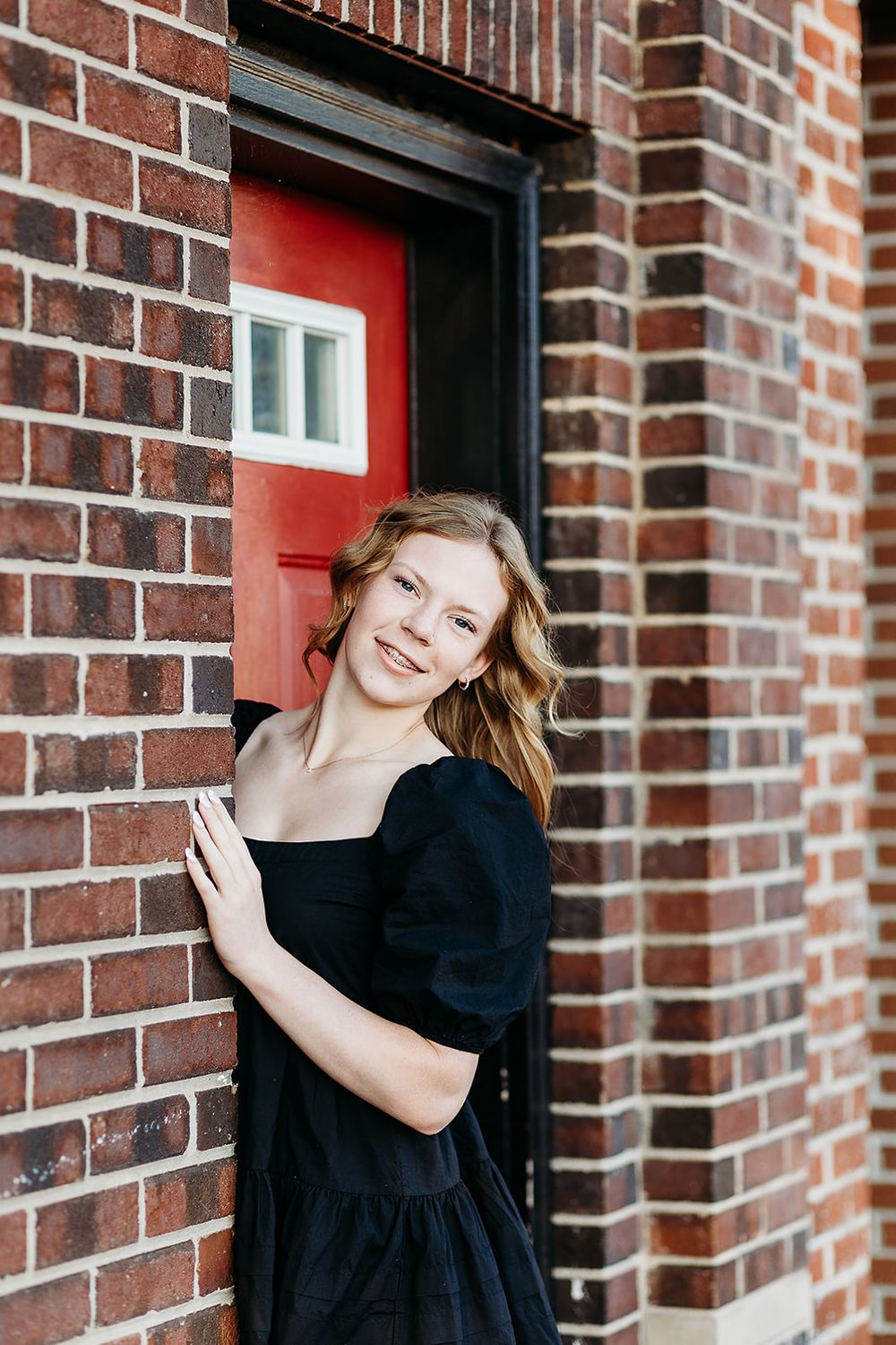 A black-dressed figure poses against a brick wall with a red door in the background.