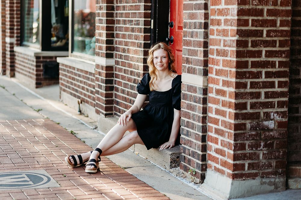 Someone in a black summer dress sits casually on a brick sidewalk against a rustic building wall.
