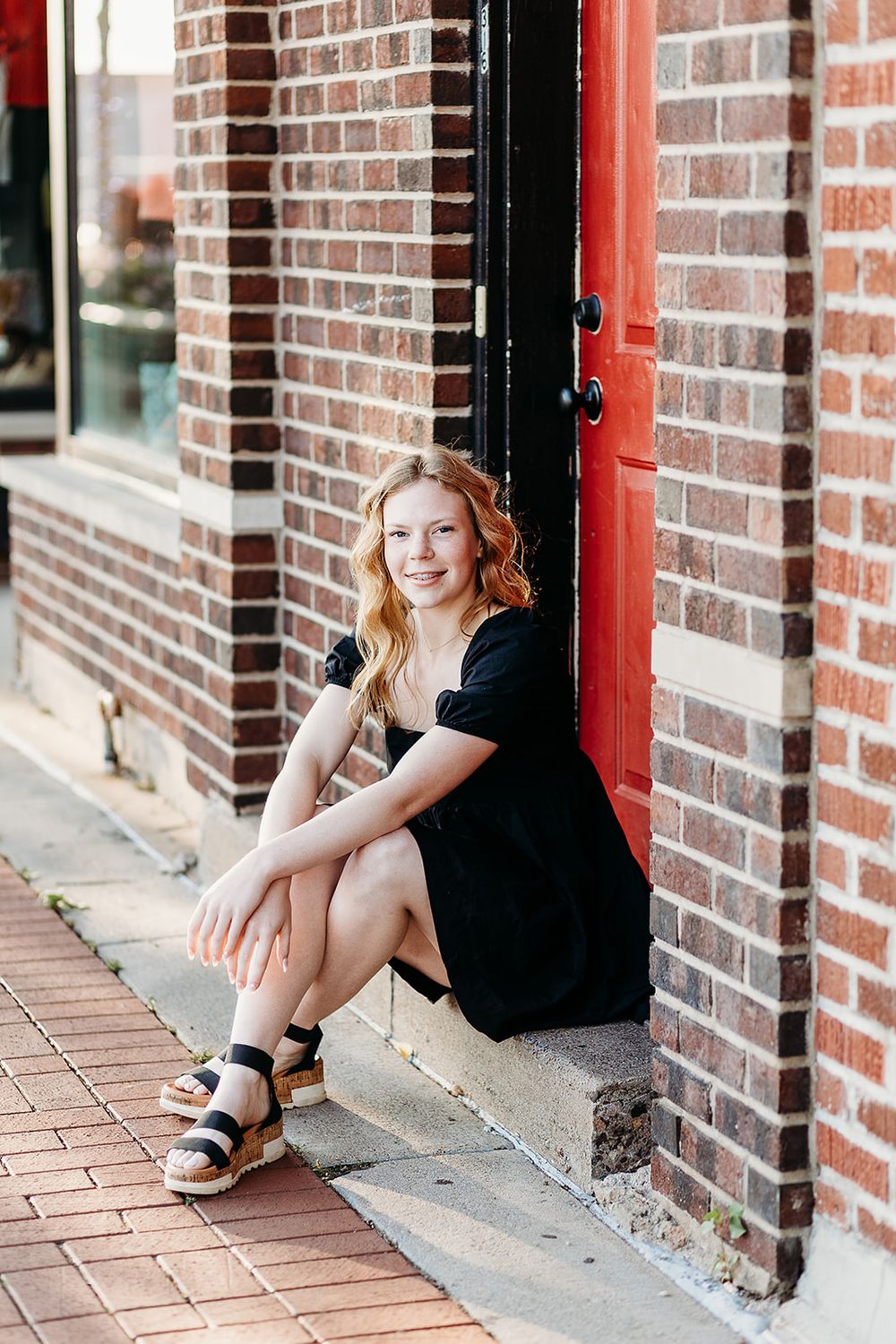 Someone in a black dress sits on brick building steps while wearing platform sandals on a sunny day.
