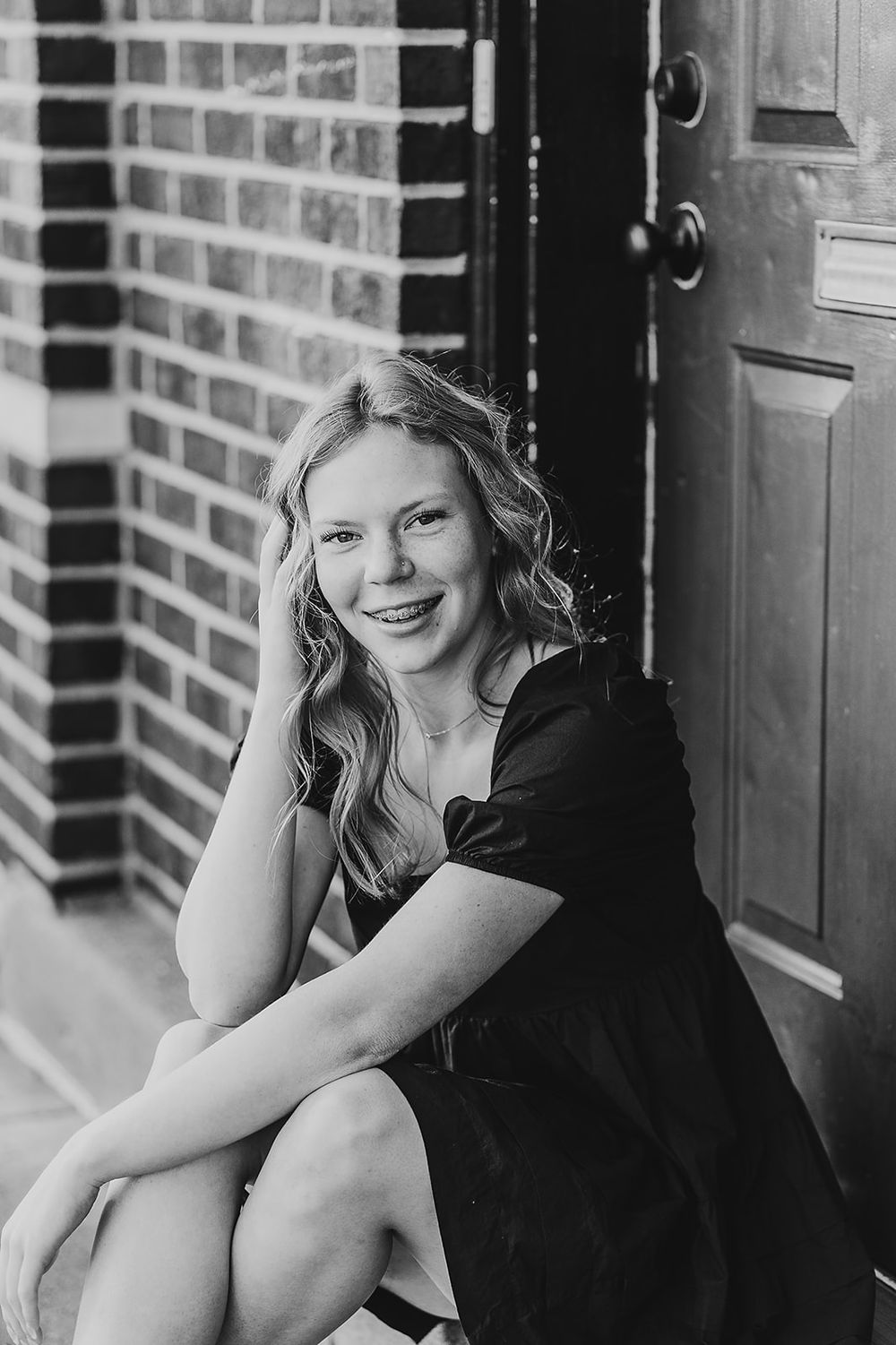 Black and white portrait of someone in a dark dress sitting by a brick wall and wooden door, smiling.