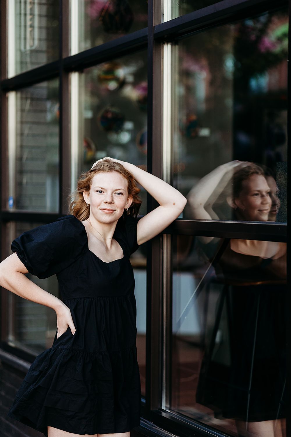 Person in black dress poses by reflective window with artful reflection creating a double image effect on city street.
