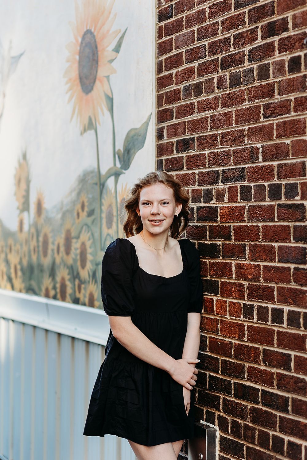 A person poses in a black dress against a brick wall with a sunflower mural in the background.