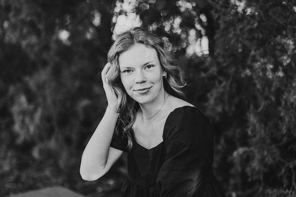 Black and white outdoor portrait of a person in a black top with wavy hair smiling against a blurred natural background.
