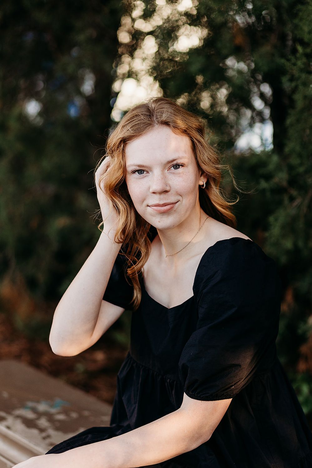 Person in black dress sitting outdoors with bokeh lights and greenery in background.