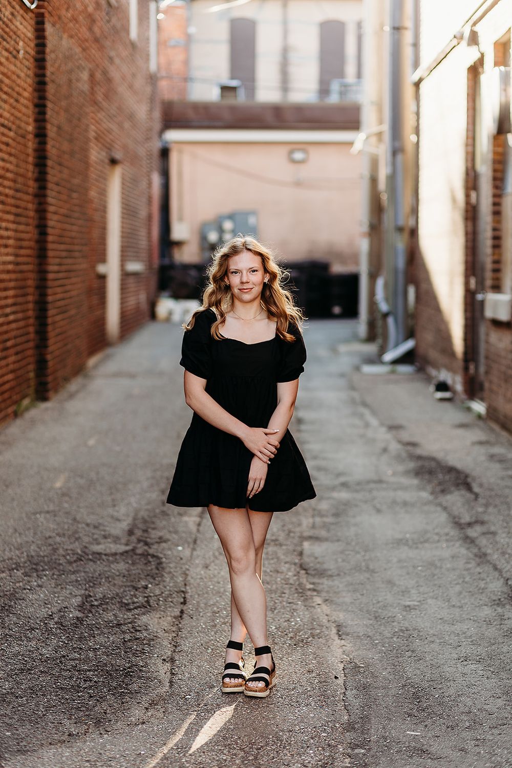 A figure in a black dress poses in an urban alleyway between brick buildings, wearing platform sandals.