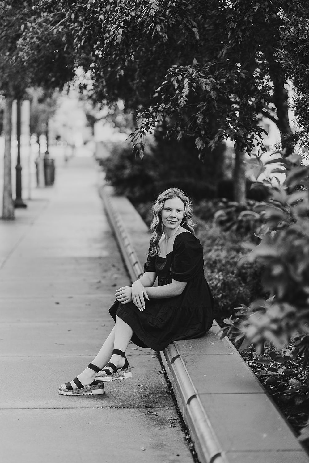 A person in a black dress and sandals sits casually on a concrete ledge along a tree-lined sidewalk in black and white.