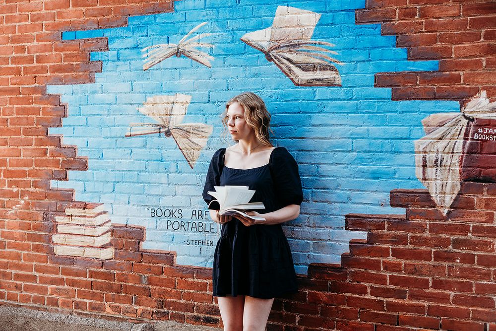 Someone in a black dress stands against a brick wall with a painted blue sky and flying paper artwork mural.