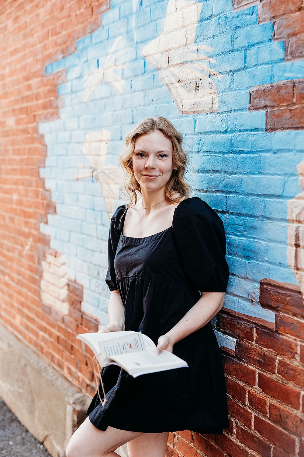 Person in a black dress stands against a brick wall painted with blue and orange colors while holding a book.
