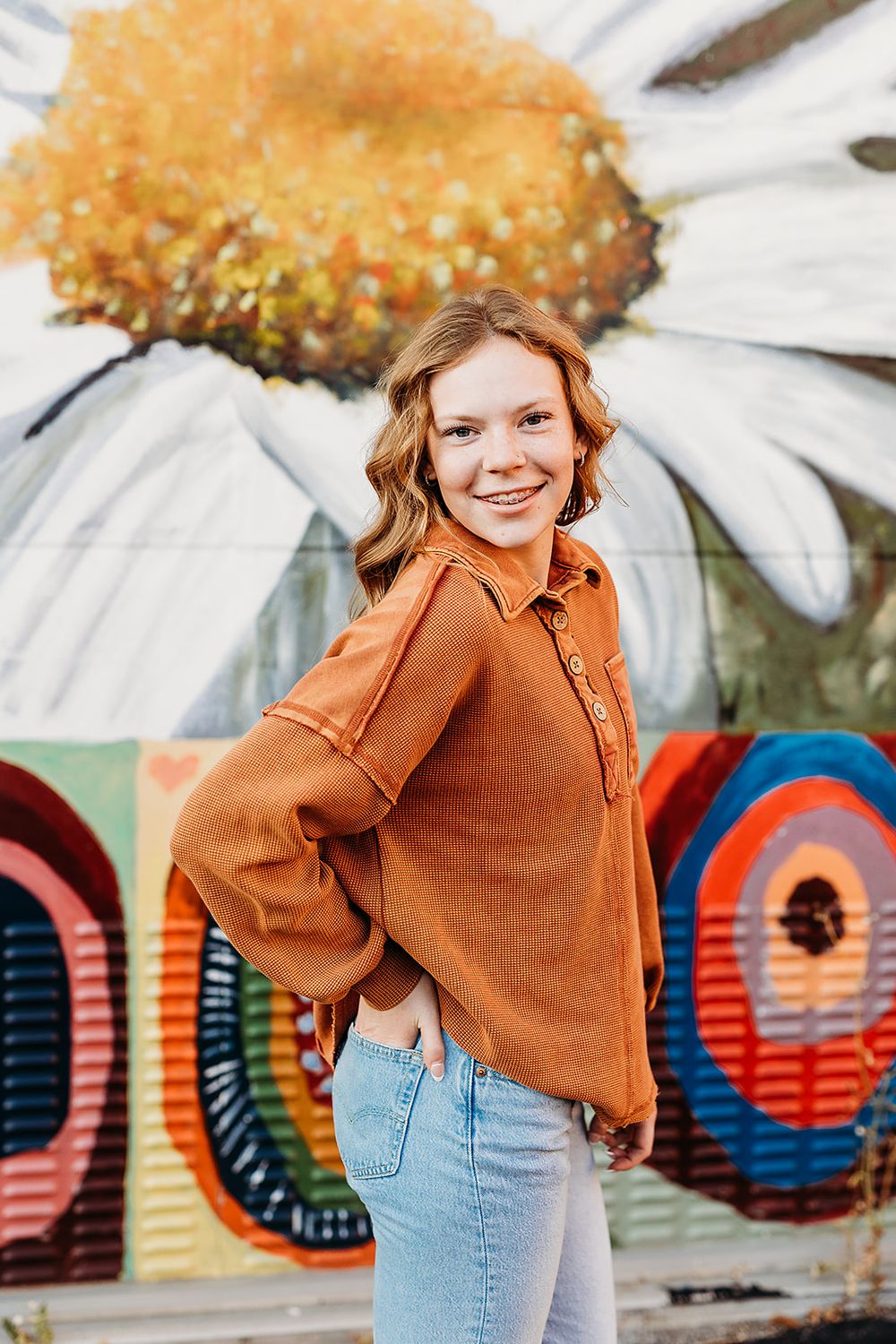 Person in rust colored sweater poses in front of colorful urban mural with daisy flower design.