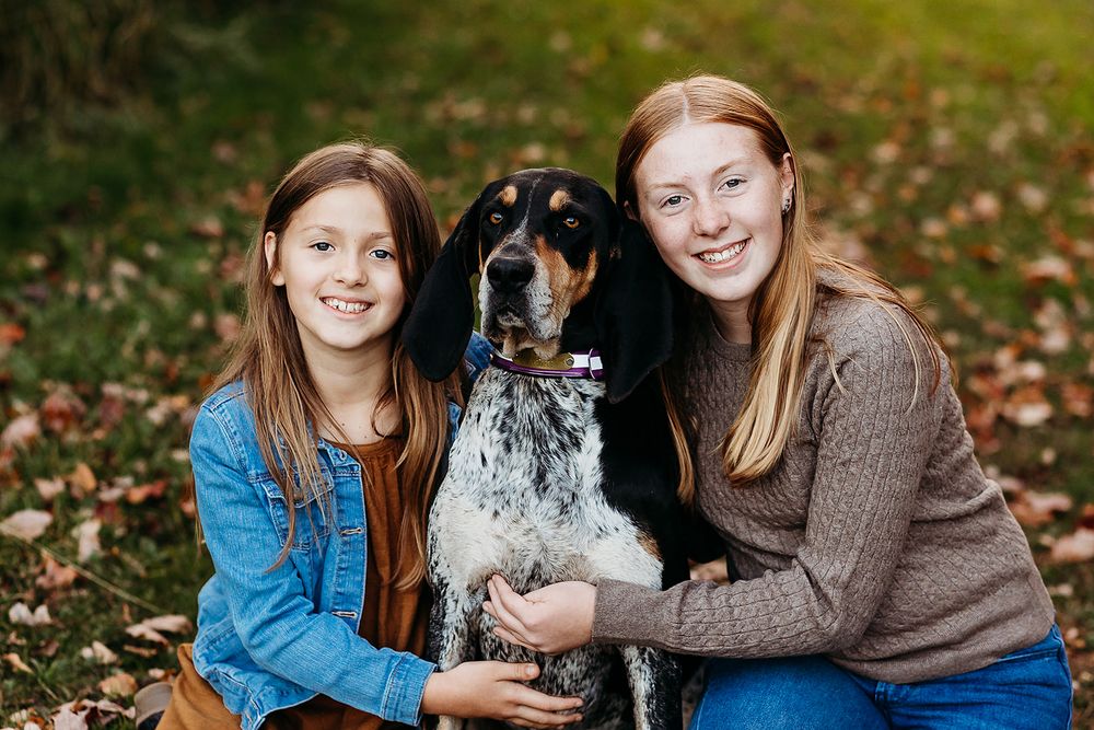 A happy Bluetick Coonhound dog sits in autumn leaves with two companions in casual sweaters during an outdoor photo session.