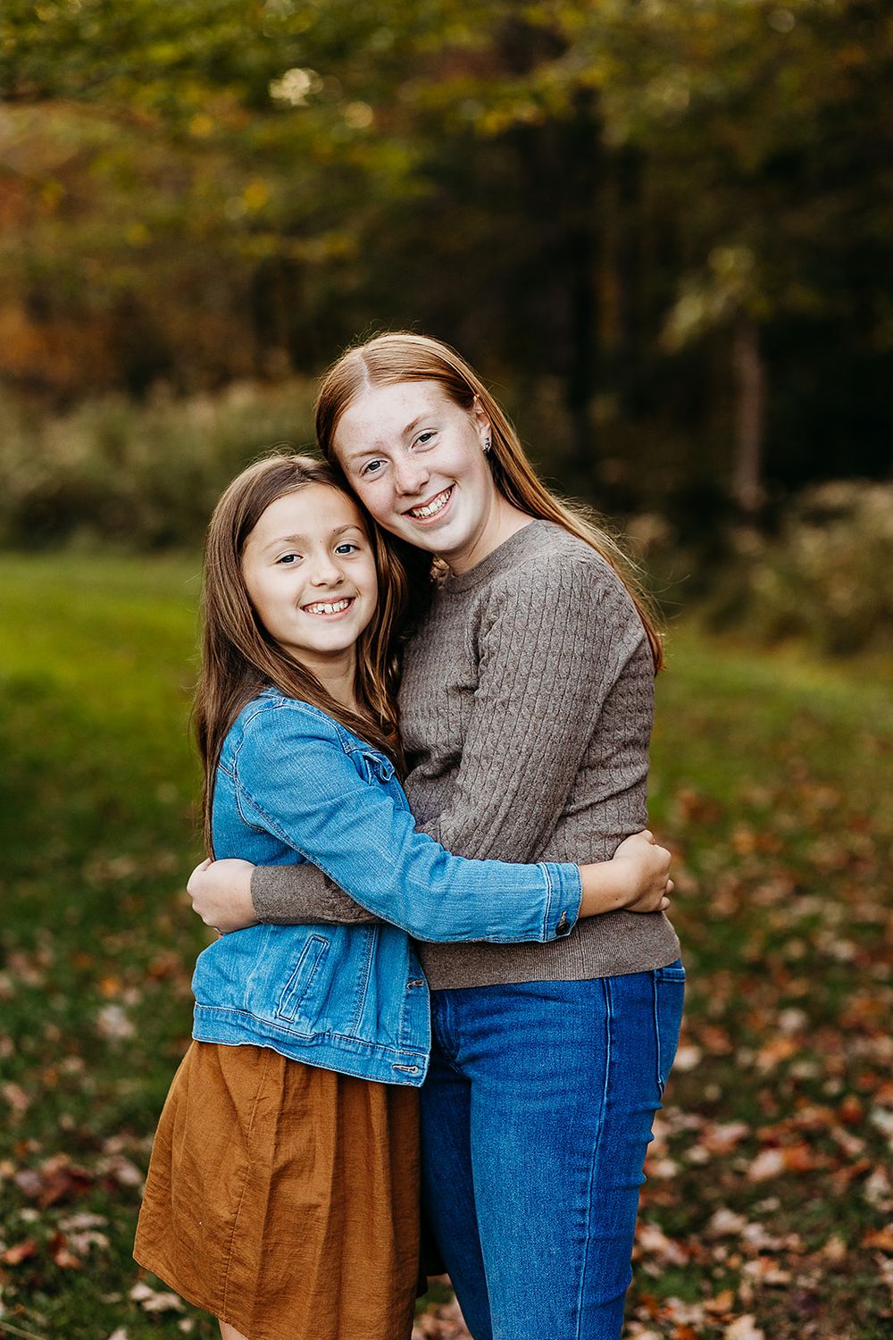 Two people embrace outdoors in autumn with fallen leaves on the ground while wearing casual clothing.