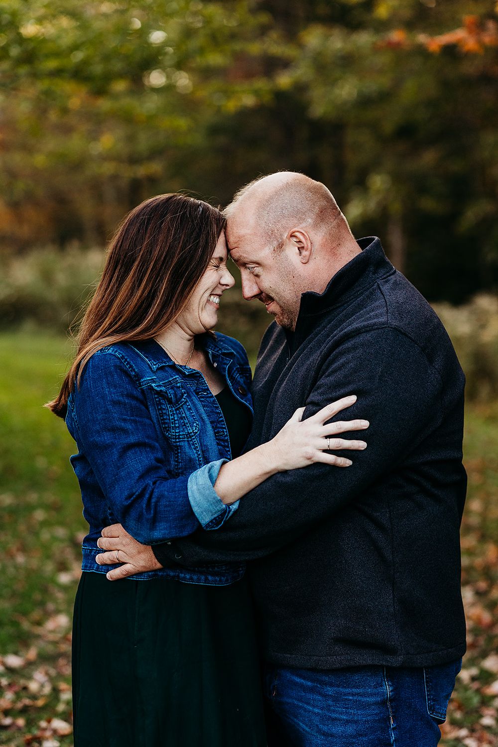 A couple laughs together in an intimate moment outdoors during autumn, wearing casual denim and dark clothing.